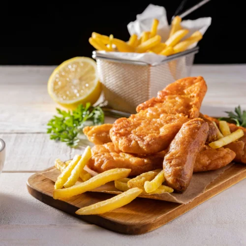 Gluten-free fish and chips served with parsley, lemon, tartar sauce, and a cold drink on a wooden table.